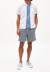 Camicia bianca a maniche corte con motivo blu, t-shirt blu sotto, pantaloni corti a righe e scarpe slip-on azzurre. Abbigliamento estivo casual.