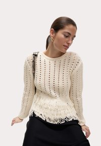 Sweter dziany w kolorze kremowym z otwartym wzorem, długimi rękawami i frędzlami na dolnym brzegu. Noszony na czarnej spódnicy, podkreślony małą torebką na pasku.
