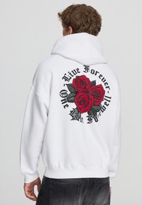 Felpa bianca con cappuccio, con una grafica di tre rose rosse e testo sul retro. Polsini e orlo a coste, texture morbida.
