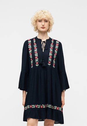 Kvinde med krøllet blondt hår iført en sort kjole med lodrette og vandrette røde blomster- og grønne bladmønstre, stående foran en ensfarvet baggrund.