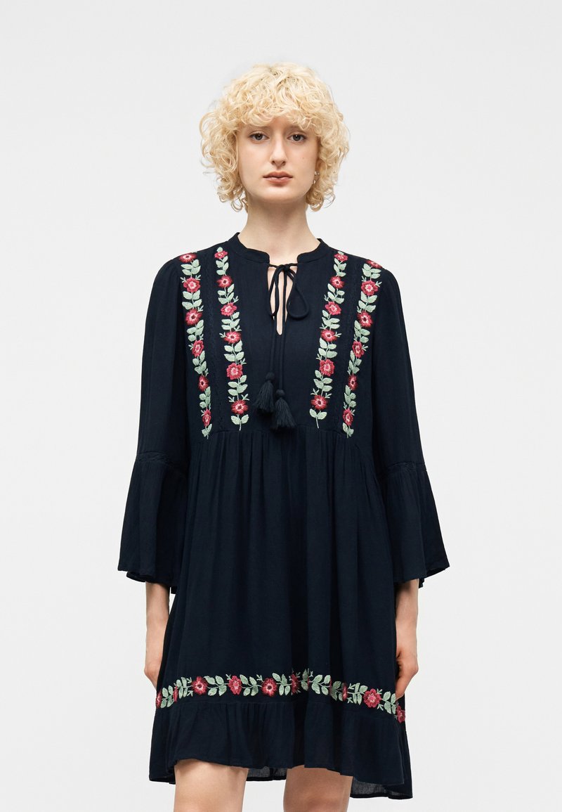 Γυναίκα με ξανθά, σγουρά μαλλιά, φορούσα μαύρο φόρεμα με κάθετη και οριζόντια κεντήματα από κόκκινα λουλούδια και πράσινα φύλλα, στέκεται μπροστά σε απλό φόντο.