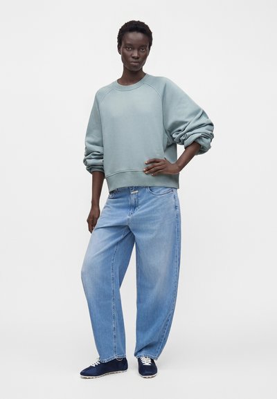 Femme aux cheveux courts portant un sweat-shirt ample bleu clair, un jean large délavé clair et des baskets bleu foncé, debout avec la main sur la hanche.