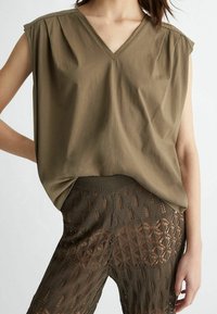 Olivově zelený top s krátkým rukávem, výstřihem do V a nařasenými rameny, doplněný průsvitnými hnědými kalhotami se vzorem.