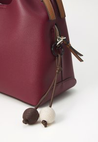 Burgunderfarbene Lederhandtasche mit strukturiertem Oberflächen, braunen Ledergriffen und dekorativen Quasten mit einem dunklen und hellen runden Perlenmuster.
