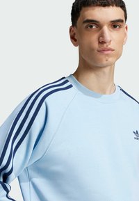 Sudadera de color azul claro con diseño de tres rayas en navy en las mangas y un pequeño logo en el pecho. Material suave con un ajuste relajado.