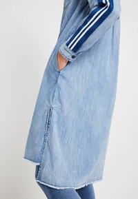 Ljusblå denimklänning med långa ärmar, navy-randiga detaljer, sidofickor och fransad fåll. Har en dragkedjedetalj fram.