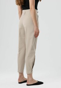 Pantalon beige à coupe ajustée, présentant une taille haute, des jambes fuselées et des boutons décoratifs à l'ourlet. Porté avec des chaussures noires sans lacets.