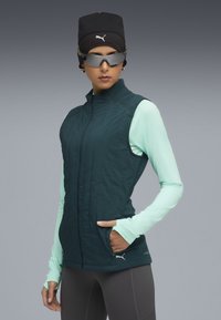 Gilet senza maniche verde scuro con colletto alto, abbinato a una maglietta a maniche lunghe di un delicato verde menta. Accentuato da un logo e leggings grigi.