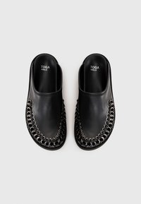 Chaussures en cuir noires à enfiler avec un détail en chaîne argentée autour des bords, semelles plates, et "TOGA VIRILIS" imprimé à l'intérieur.
