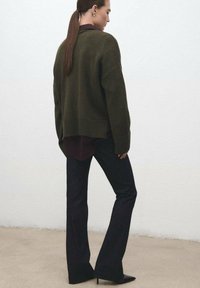 Suéter de punto oversize en verde oliva, superpuesto a una camiseta burdeos, combinado con pantalones negros acampanados y tacones negros de punta.