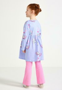 Rödahårigt barn ifört en ljusblå klänning med tecknade fjärilar och rosa hjärtan, kombinerat med knallrosa byxor och sneakers, står i profil.