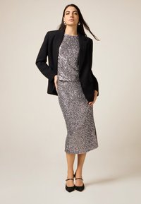 Kvinde iført en sølvfarvet paljetkjole med en sort blazer og sorte hæle, stående mod en ensfarvet beige baggrund.