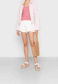 Haut bustier en vichy rouge avec basque, veste oversize rose clair, short blanc effiloché et sac tressé marron, assortis à des sandales blanches.