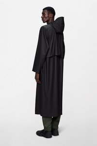 Μακρύ μαύρο αδιάβροχο με κουκούλα, ματ υφή, με κοντή σχεδίαση στη μέση και μακριά μανίκια. Φορεμένο πάνω από πράσινο παντελόνι και μαύρα παπούτσια.