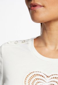 Biała bawełniana koszula z okrągłym dekoltem, ozdobnymi wycięciami w spiralnym wzorze oraz srebrnymi guzikami na ramieniu.