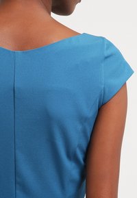 Person, die ein kurzärmliges blaues Oberteil trägt, von hinten und über die Schulter gesehen, vor einem einfarbigen hellen Hintergrund.