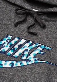 Grå hættetrøje med et fremtrædende blåt og hvidt tie-dye Nike-logo. Sorte snore supplerer det teksturerede stof.