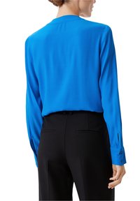 Blusa a maniche lunghe di un blu vivace, realizzata in tessuto liscio. Presenta un collo rotondo e polsini con bottoni. Abbinata a pantaloni neri dal taglio sartoriale.