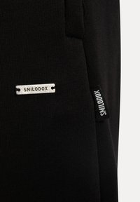 Czarne spodnie sportowe z prążkowaną strukturą, z metalową tabliczką z logo "SMILODOX" w kolorze srebrnym oraz małą czarną metką po boku.