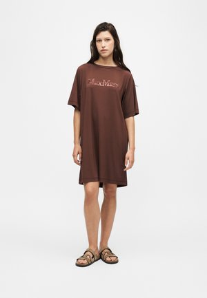 Femme portant une robe T-shirt midi marron fluide avec le logo "MaxMara", associée à des sandales marron clair à double bride, debout sur un fond uni.