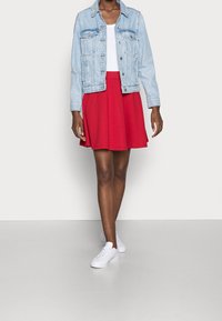 Veste en denim bleu clair, t-shirt blanc, jupe évasée rouge et baskets blanches. Comprend des poches et un design court.
