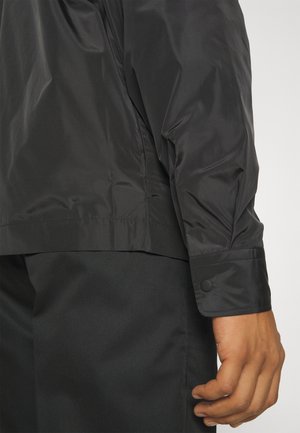 Plano detalle del brazo de una persona que lleva una camisa de manga larga negra con puños abotonados y pantalones negros, con un fondo liso.