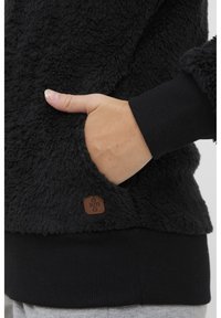 Sort, fluffy fleece-top med en lomme på siden, der har en ribbet sort manchet og et lille brunt logo-tag i kanten.