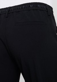 Pantalon noir avec une ceinture élastique extensible froncée, une poche arrière unique et une texture de tissu lisse. Design minimaliste.