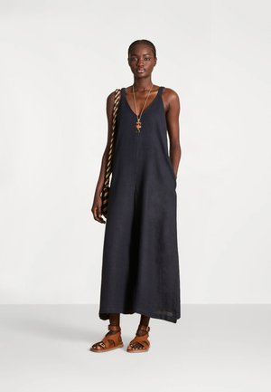Femme portant une combinaison noire sans manches à jambes larges, des sandales marron, un long collier et un sac à bandoulière rayé sur un fond uni.