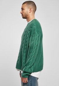 Green cable-knit sweater with a relaxed fit, textured pattern, and ribbed cuffs and hem. Worn over a white T-shirt and denim jeans.