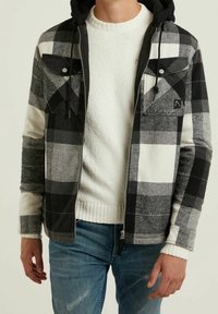 Czarno-biała kraciasta kurtka z kapturem, zapinana na zamek, z dwoma przednimi kieszeniami. Noszona na białym, fakturowanym swetrze. Widoczne niebieskie jeansy.