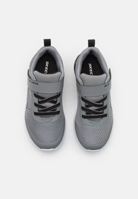 Baskets de sport grises avec une tige en mesh, lacets noirs et une sangle au milieu du pied. Semelle épaisse blanche avec une finition texturée.