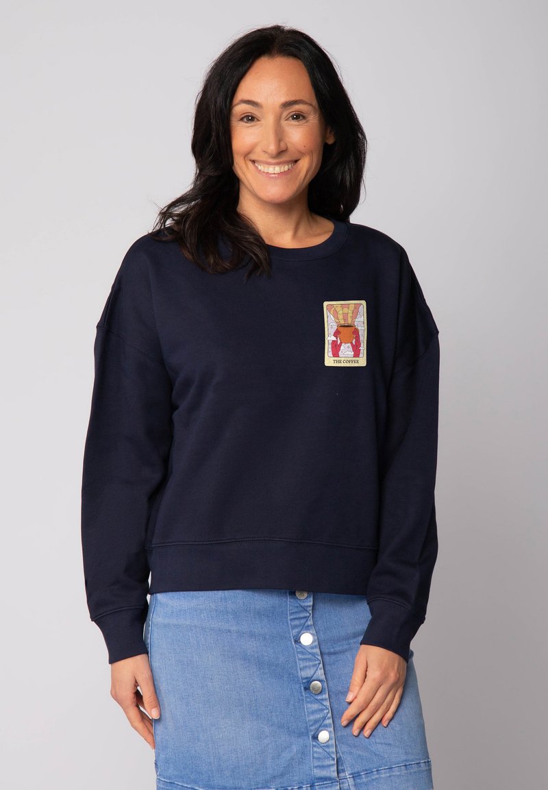 Femme souriante aux cheveux foncés portant un sweat-shirt bleu marine avec un patch "The Coffee" et une jupe en denim bleu clair à boutons, se tenant devant un fond uni.