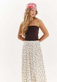 Žena s dlhými blond vlasmi, oblečená v červenej kvetinovej šatke, čiernom topu bez ramienok, náhrdelníku z korálkov a bielej sukni s čiernymi bodkami.