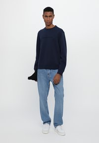 Joven vestido con sudadera azul marino, jeans de mezclilla claros, zapatillas blancas, sosteniendo una gorra negra, de pie frente a un fondo blanco liso.