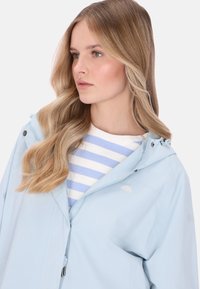 Világoskék vízálló kabát kapucnival, cipzáros lezárással, húzózsinórral és diszkrét logóval, csíkos felső fölött viselve.