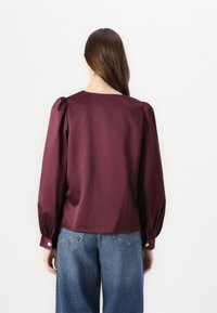 Burgundy blouse with long puff sleeves, smooth texture, and a wide neckline. Paired with loose blue denim jeans.
