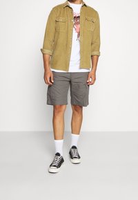 Camisa de pana marrón, camiseta gráfica blanca, pantalones cortos de cargo grises, calcetines tobilleros blancos, zapatillas negras. Atuendo casual con diversas texturas.