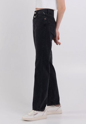 Persoon die zwarte high-waist jeans met rechte pijpen, een witte cropped top en witte sneakers draagt, zijwaarts staand op een effen lichte achtergrond.