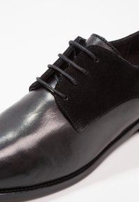 Chaussure habillée en cuir noir avec une finition lisse, des accents en daim et six œillets pour les lacets. Bout arrondi et bords de semelle surpiqués.