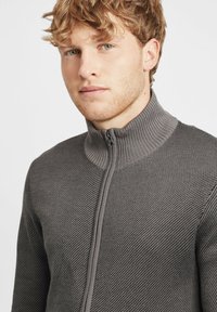 Suéter de cuello alto con cremallera en punto gris texturizado. Presenta un cuello y puños acanalados, diseño ajustado y detalles de costura lisa.