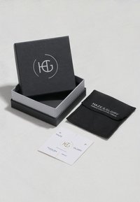 Black jewelry box with silver logo, gray interior, black fabric pouch, and a white care card with gold accents, all arranged neatly.