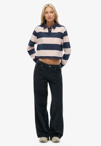 Vrouw staat met het gezicht naar voren in een cropped langemouwen polo shirt met marineblauwe en crème gestreepte patronen, zwarte wijde broek en beige sneakers.