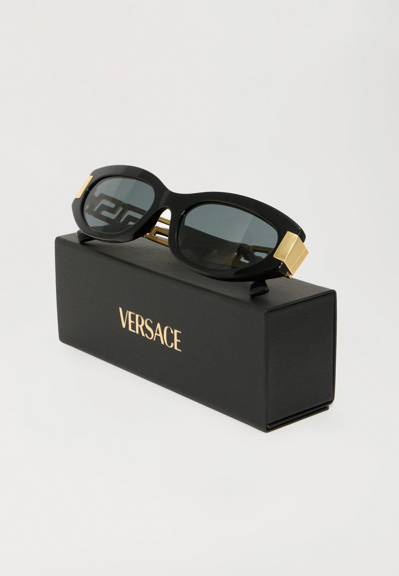 Zwarte oversized zonnebril met grijs getinte lenzen, gouden accenten en gedurfde, rechthoekige montuur, geplaatst op een zwarte doos met het label "Versace."