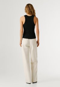 Femeie cu păr ondulat castaniu deschis, purtând un maiou negru mulat și pantaloni largi de culoare alb-crem, stând cu fața spre departe pe un fundal simplu.