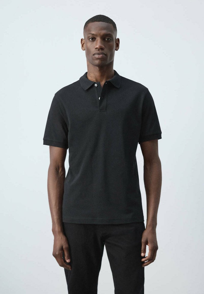 Homme debout sur un fond uni, portant un polo noir à manches courtes et un pantalon noir, face à la caméra avec une expression neutre.