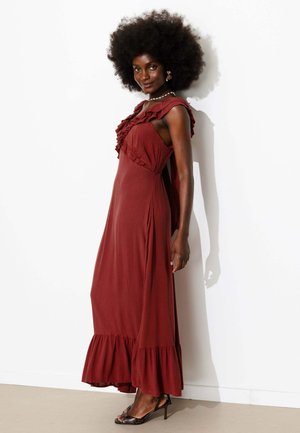 Femme avec une grande coiffure afro portant une robe longue sans manches rouille à volants et des sandales noires à talons, debout contre un mur blanc.