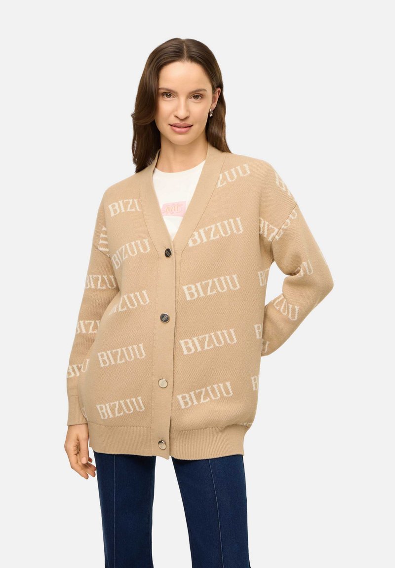 Vrouw draagt een beige oversized cardigan met het tekstpatroon "BIZUU", gecombineerd met donkerblauwe jeans en een wit topje, staand met één hand in de zij.