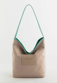 Beige syntetisk väska med turkos foder och rem, som har en stor öppning och en beige präglad etikett på framsidan.