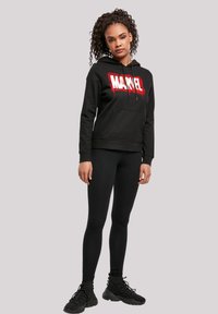 Felpa nera con logo "MARVEL" rosso e bianco, abbinata a leggings neri e scarpe da ginnastica nere, adatta per un abbigliamento casual.